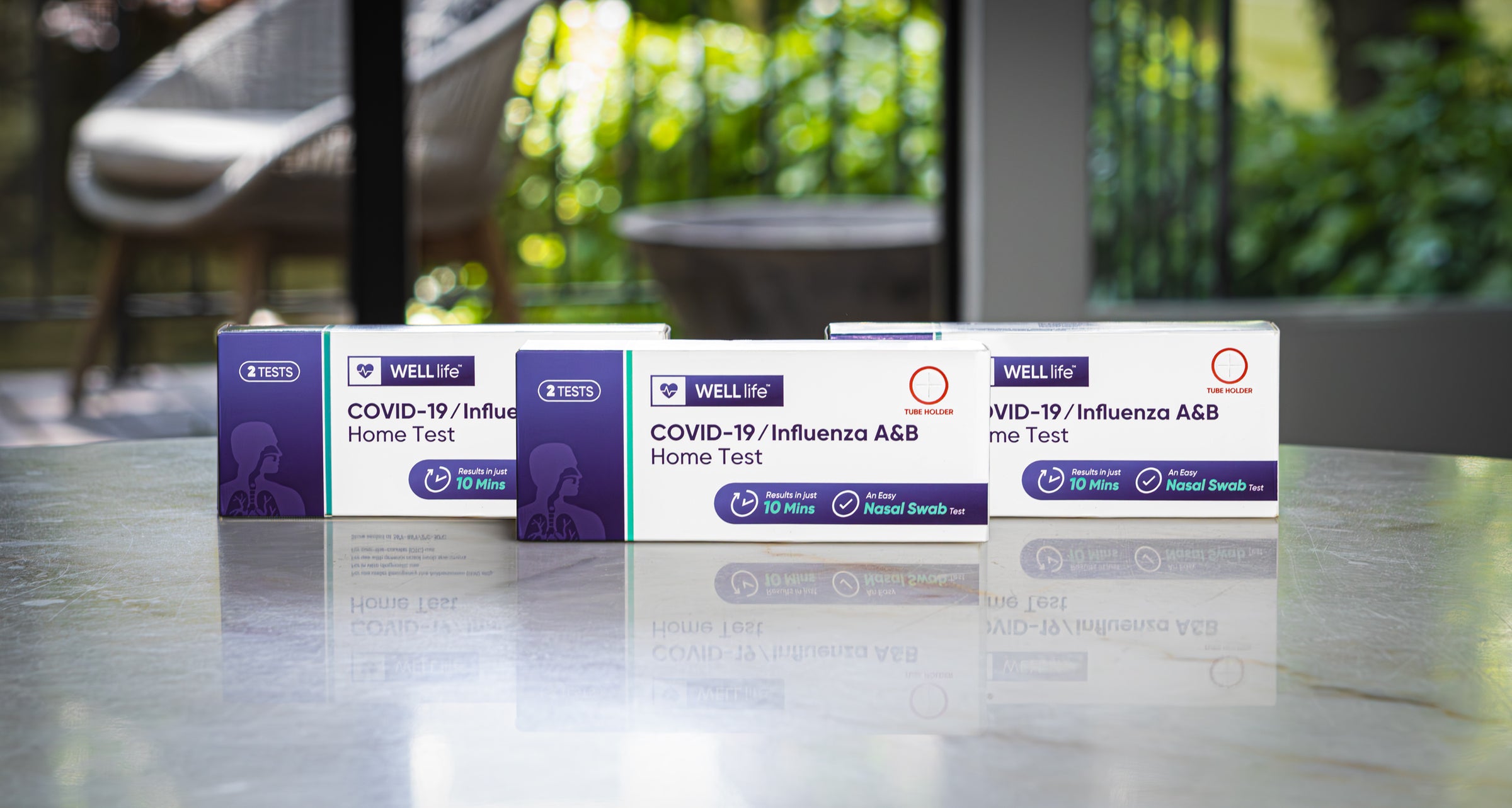 Three WELL life™ COVID-19 & Influenza A & B Rapid at Home Test Kits on a table in front of a large window